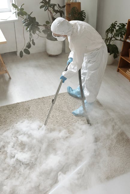 A woman with blonde hair tied back, dressed in a beige jacket, white top, and jeans, vacuuming a patterned area rug in a living room. The rug features a floral design in shades of beige, orange, and b