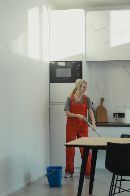 A woman with blonde hair wearing a red apron and grey shirt is mopping the light wooden floor of a modern kitchen, which features white walls, a built-in microwave oven, and a marble-effect backsplash